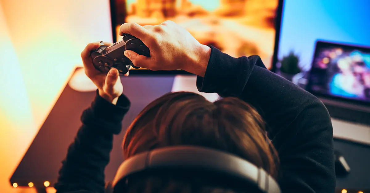 Vista desde atrás de una persona con auriculares jugando videojuegos con un controlador frente a un monitor.