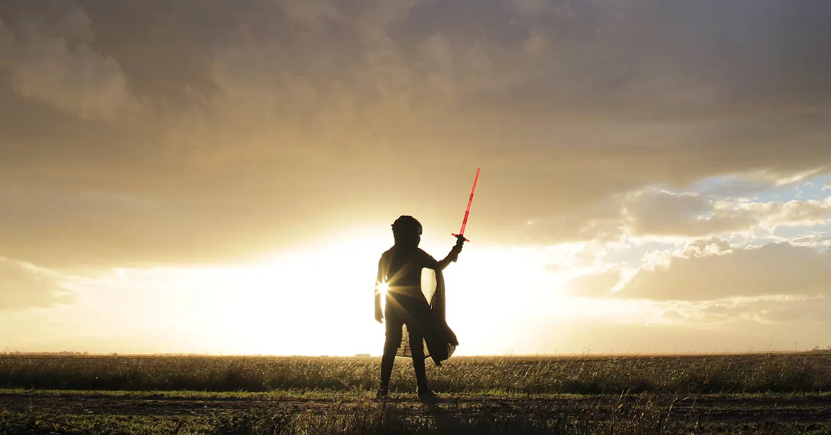 Silueta de Darth Maul sosteniendo una espada de luz roja frente a un atardecer en un campo abierto.