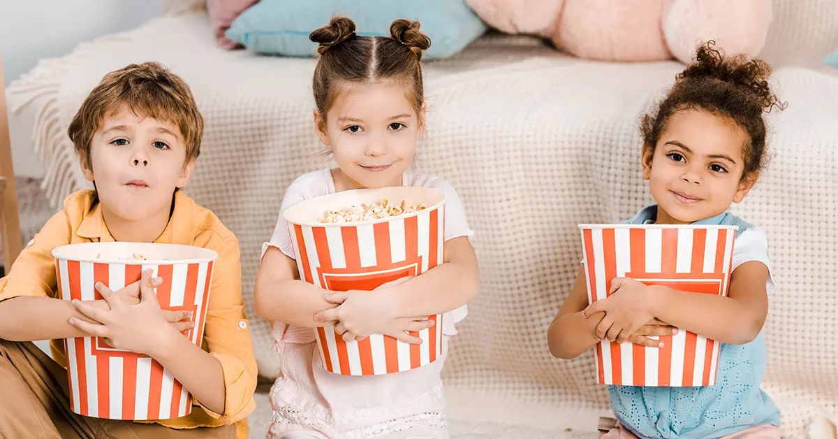 Dos niñas y un niño sentados en el suelo sosteniendo grandes botes de palomitas de maíz.